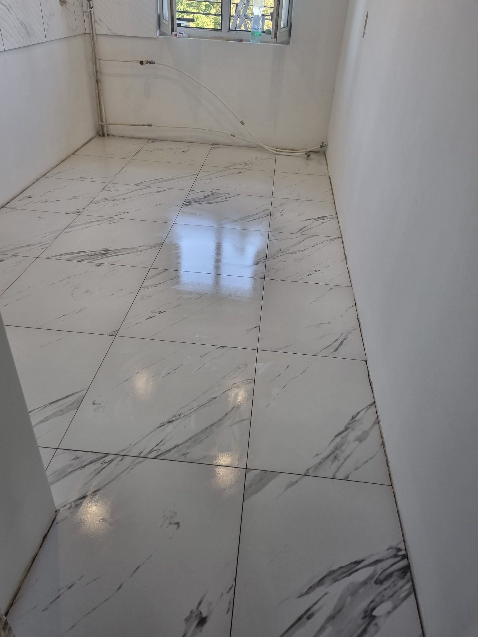 Ułożone, błyszczące płytki podłogowe imitujące marmur w jasnym pomieszczeniu z białymi ścianami i oknem w tle, widoczne rury przy ścianie.