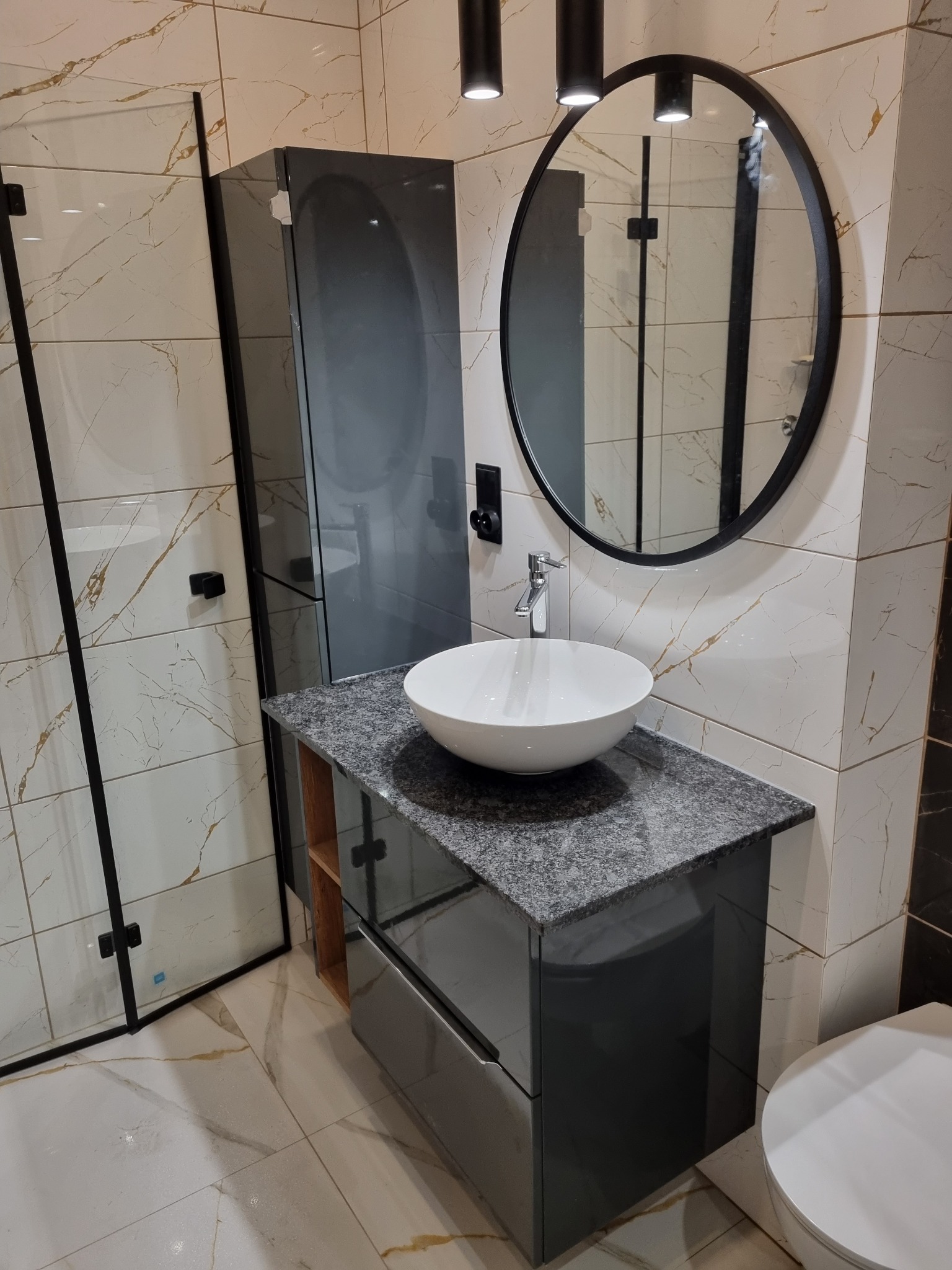 Nowoczesna łazienka z kabiną prysznicową z czarnymi ramami, umywalką nablatową na szafce z szarym blatem imitującym kamień, okrągłym lustrem w czarnej ramie i marmurowymi płytkami z złotymi żyłami.