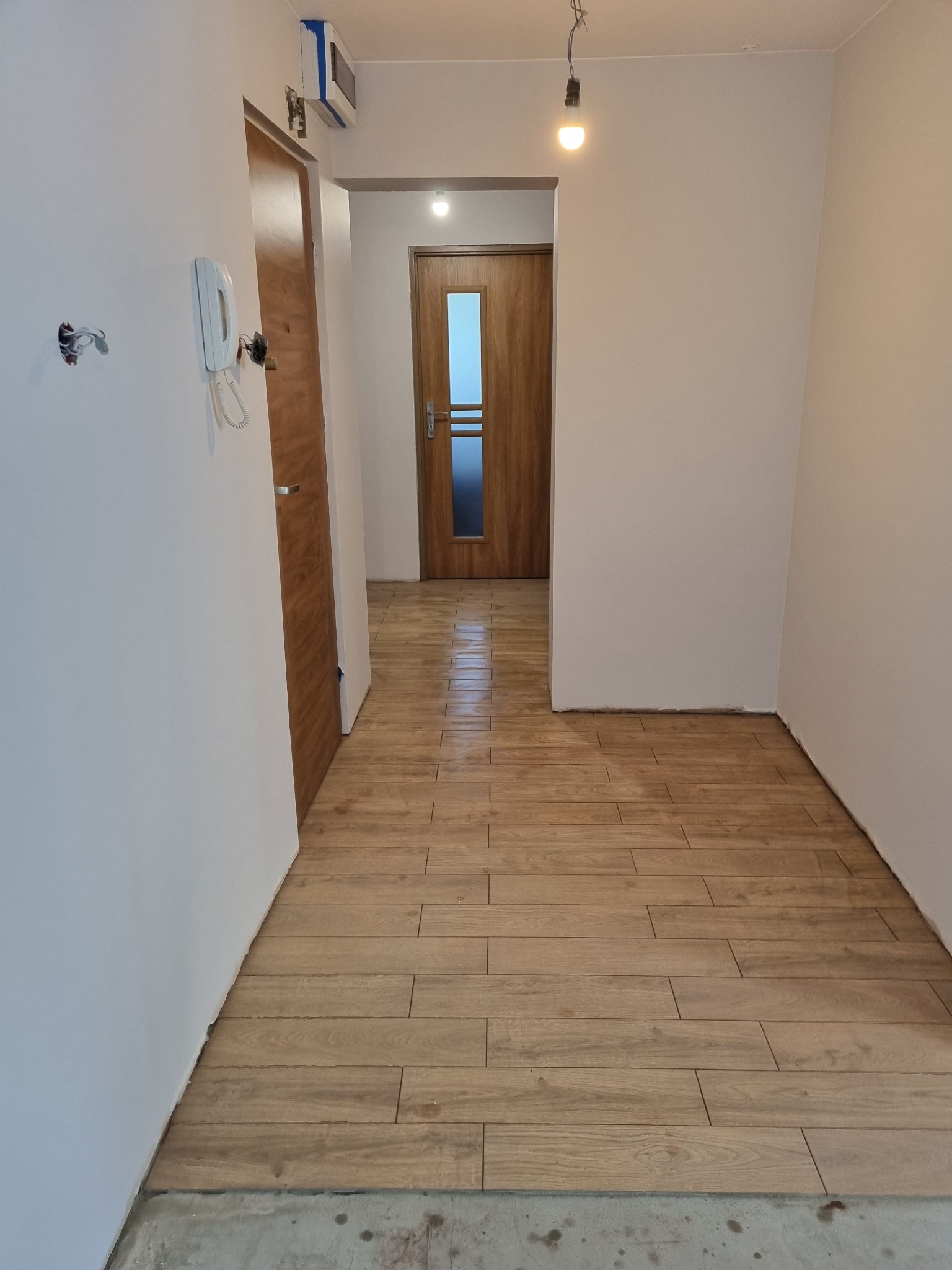 Korytarz w trakcie prac wykończeniowych: ułożona podłoga z paneli imitujących drewno, surowe ściany pomalowane na biało, widoczne niedokończone gniazdka elektryczne i wystające kable, w tle...