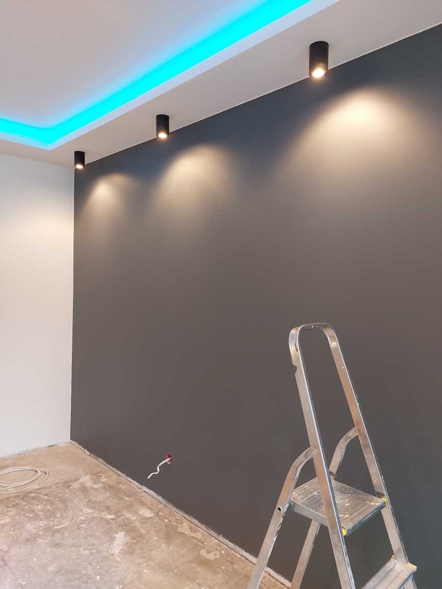 Pokój z szarą ścianą oświetloną trzema punktowymi lampami sufitowymi i niebieskim oświetleniem LED wzdłuż sufitu, widoczna aluminiowa drabina i przewody elektryczne.