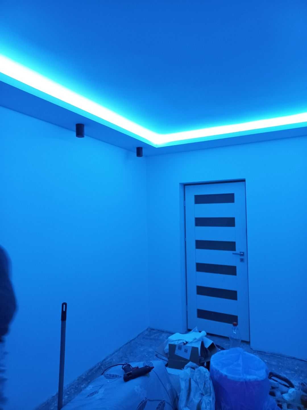 Pokój w trakcie prac wykończeniowych z zamontowanym oświetleniem LED na suficie, widoczne narzędzia i materiały budowlane na podłodze, ściany pomalowane na niebiesko.