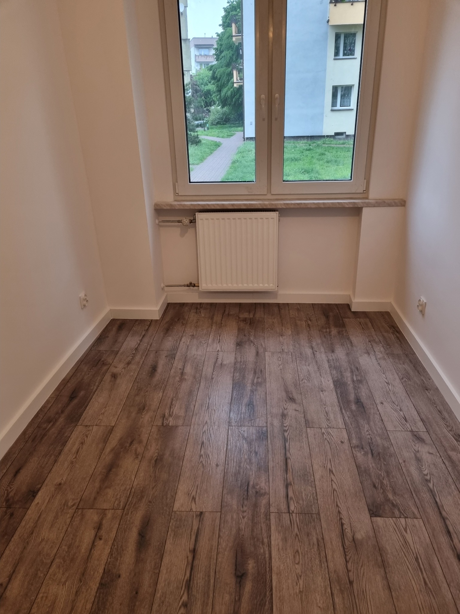 Pusty pokój po remoncie z panelami podłogowymi w odcieniach brązu, białymi ścianami i grzejnikiem pod oknem z widokiem na budynek mieszkalny.