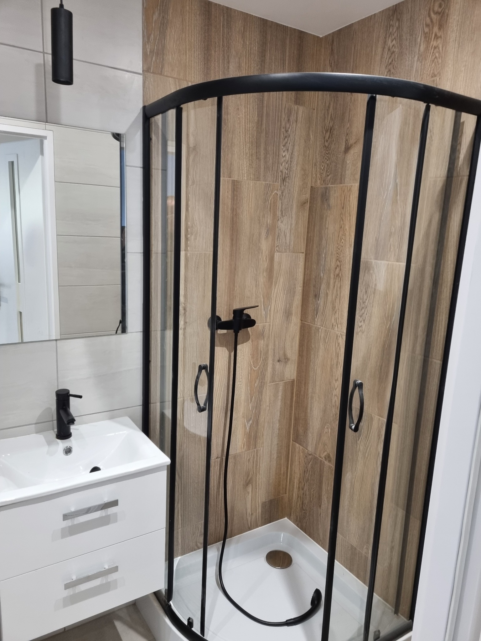 Nowoczesna łazienka po remoncie: kabina prysznicowa z czarną ramą i armaturą, umywalka z szafką w kolorze białym, drewnopodobne płytki na ścianach kabiny.
