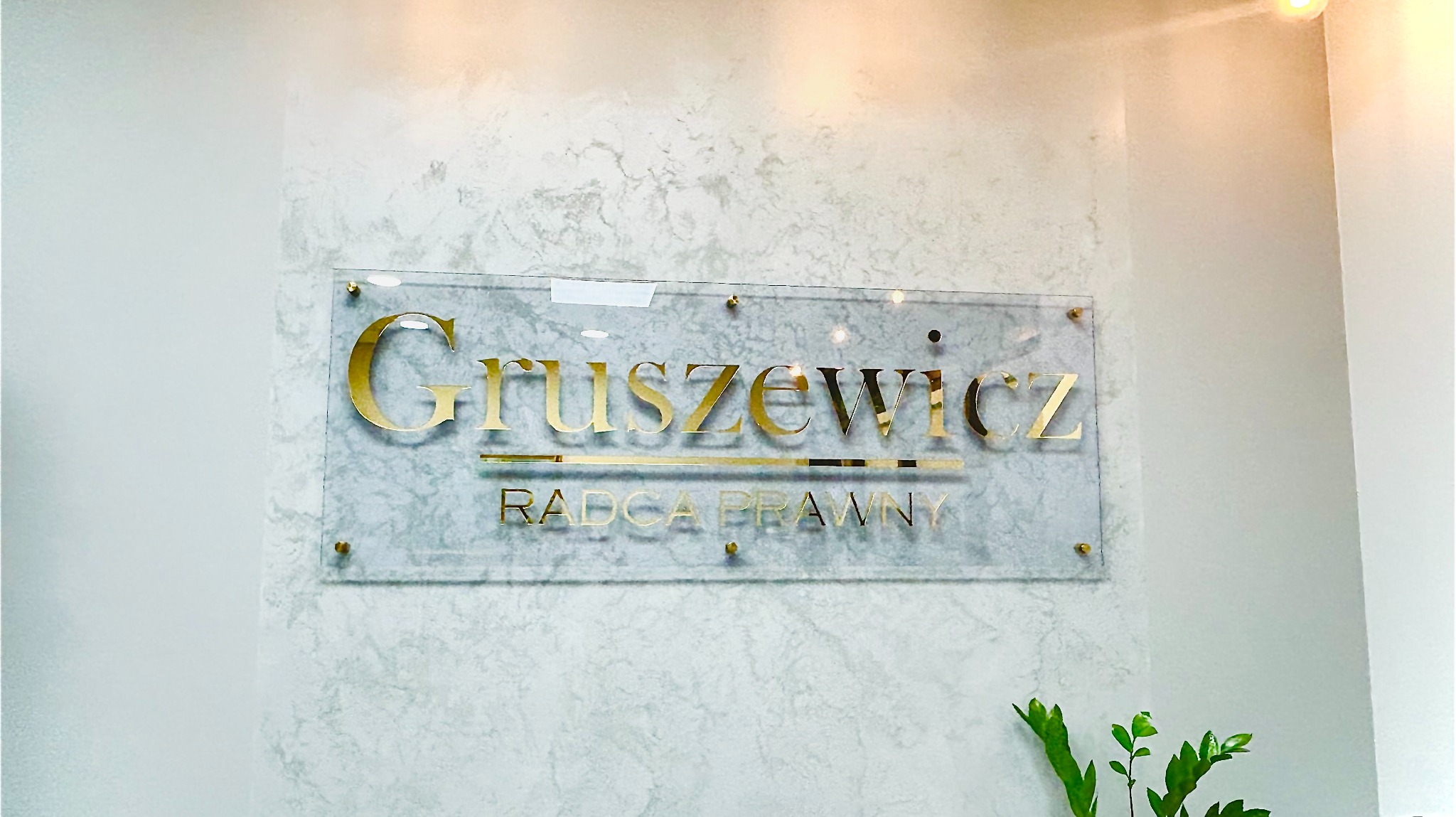 Tabliczka z nazwiskiem 'Gruszewicz' i napisem 'Radca Prawny' w złotym kolorze, umieszczona na ścianie o marmurkowej fakturze, z widocznym fragmentem rośliny w dolnym rogu.