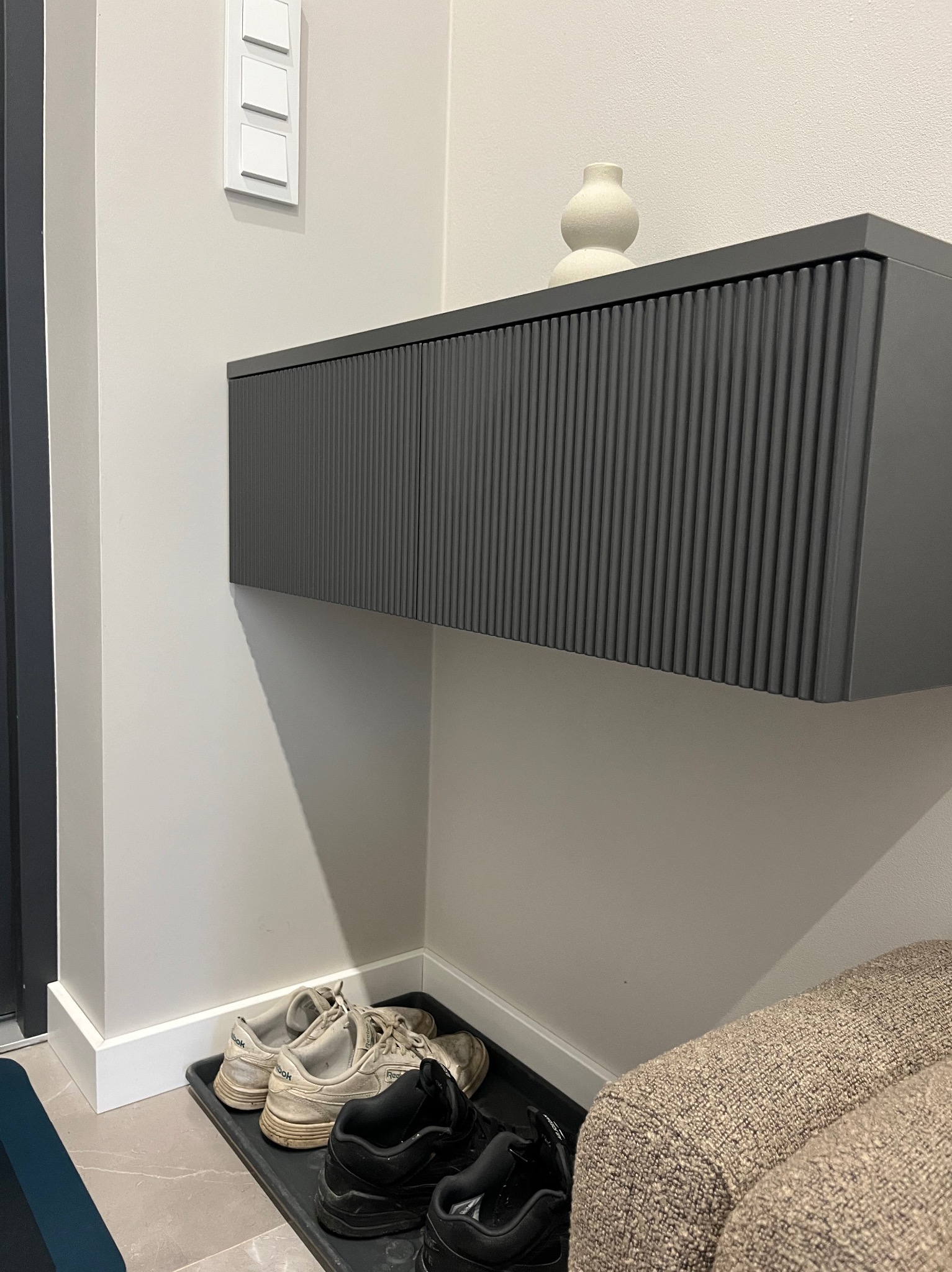 Szara, wisząca szafka z ryflowanym frontem i dekoracyjnym wazonem na górze, umieszczona w przedpokoju nad tacą z butami. Minimalistyczny design.