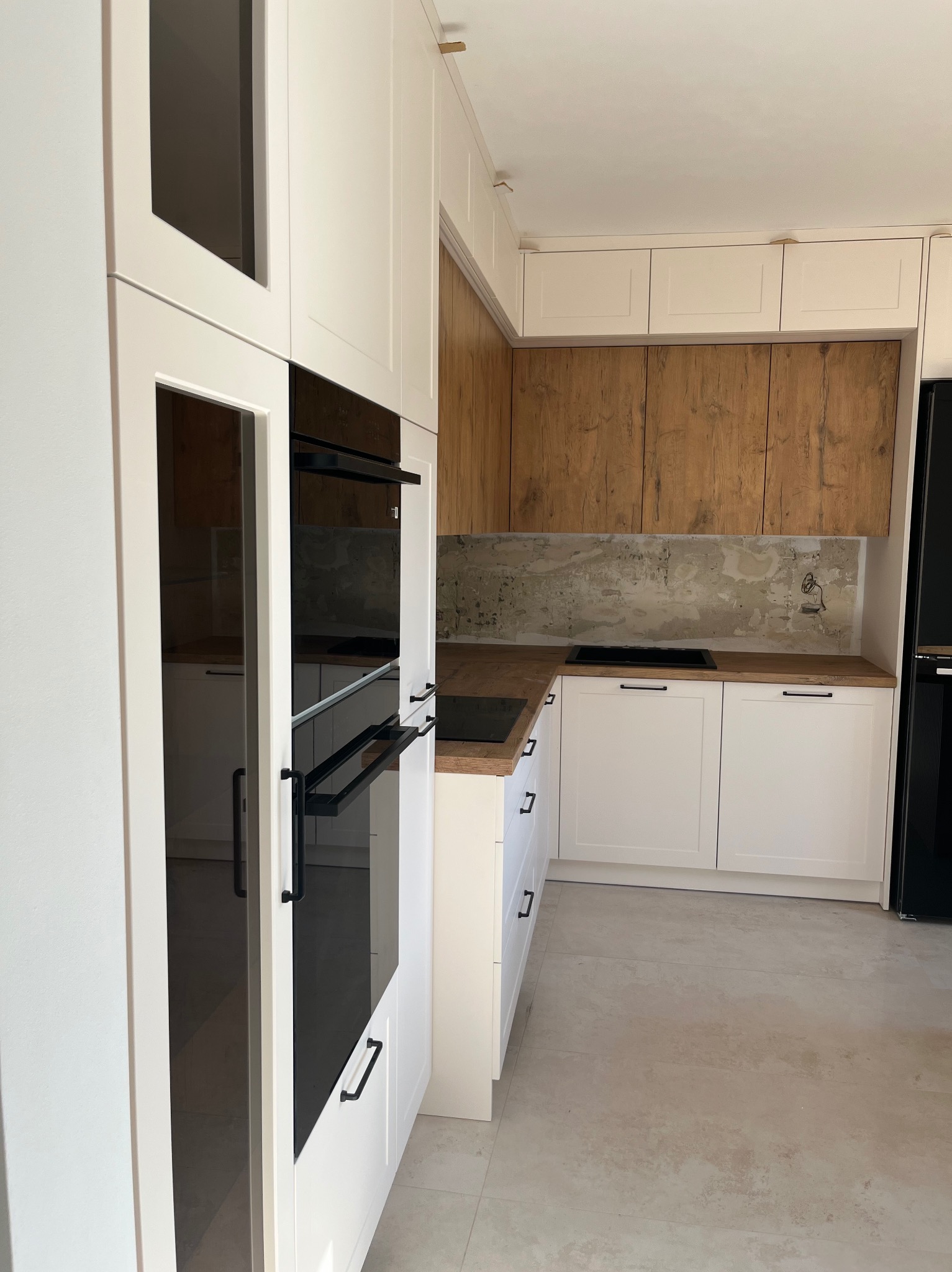 Nowoczesna kuchnia z białymi szafkami i drewnianymi akcentami. Widoczne czarne uchwyty i sprzęt AGD. Blat drewniany, ściana z surowym wykończeniem. Minimalistyczny design.