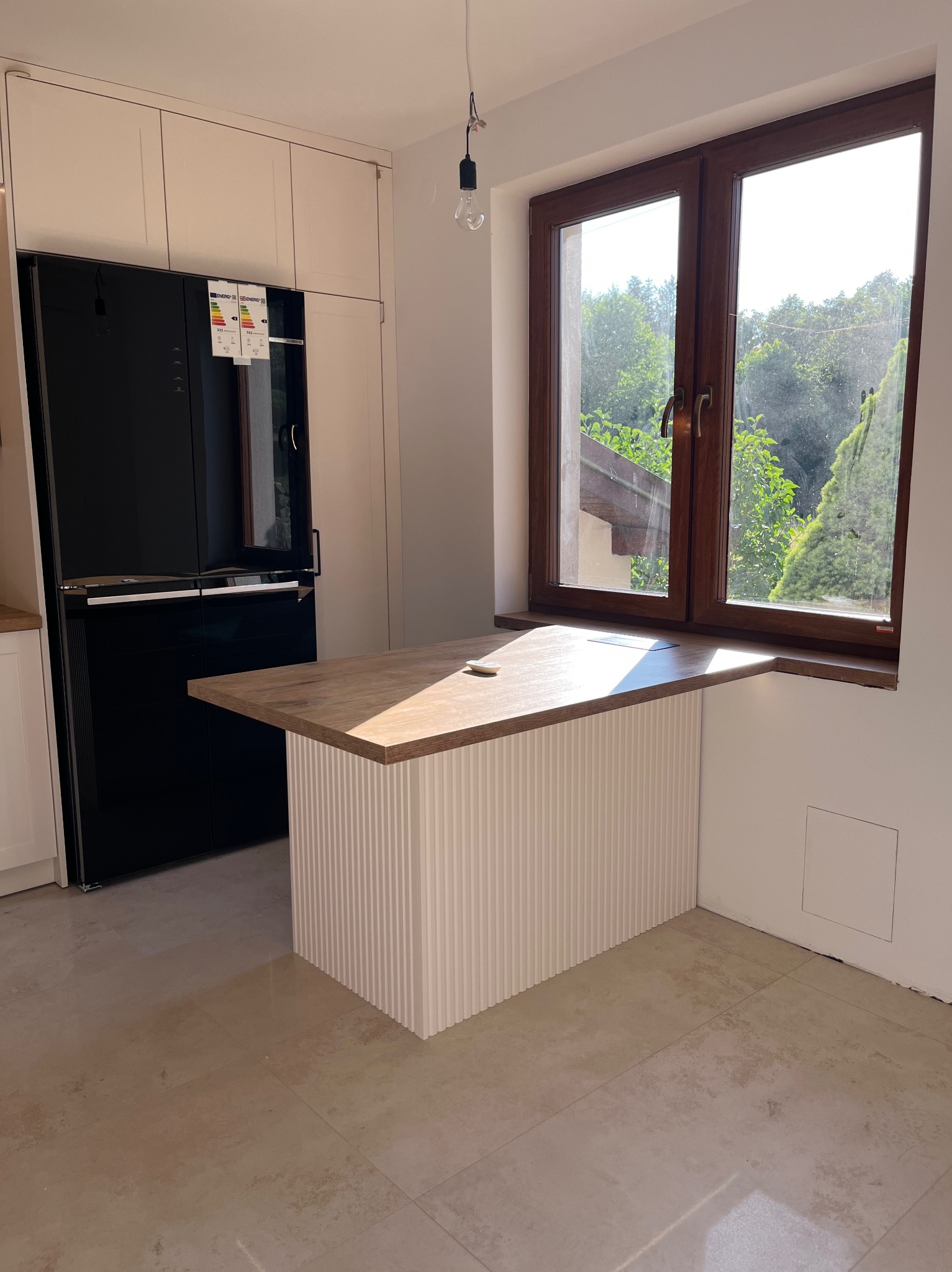 Kuchenny blat drewniany, wkomponowany w ramę okienną, z ryflowanym białym frontem. W tle czarna lodówka i jasne szafki. Widok z okna na zieleń.