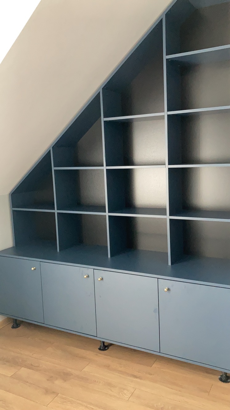 Niebieska szafa wnękowa z półkami i szafkami dolnymi, dopasowana do skosu dachu, z złotymi gałkami i regulowanymi nóżkami, na panelach podłogowych.