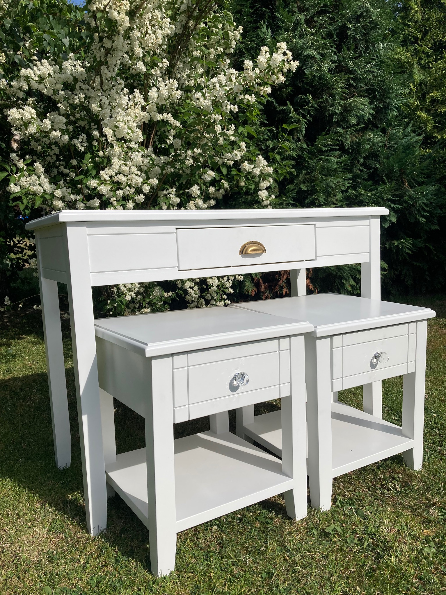 Biały, drewniany komplet mebli ogrodowych: konsola z szufladą i dwa stoliki nocne z kryształowymi gałkami, na trawie, z kwitnącym krzewem w tle.