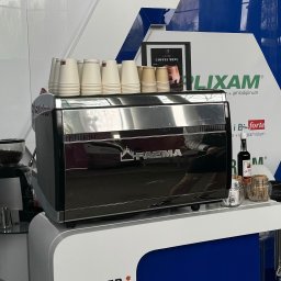Eventship Weronika Pietruszka - Profesjonalny ekspres do kawy marki Faema z zapasem papierowych kubków, młynek do kawy i dodatki na blacie w otoczeniu materiałów reklamowych.