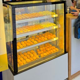 Eventship Weronika Pietruszka - Podświetlana witryna chłodnicza z trzema półkami pełnymi wypieków: makaroniki w odcieniach żółci i pomarańczy oraz małe rogaliki. W tle widoczni ludzie i elementy wystawiennicze.