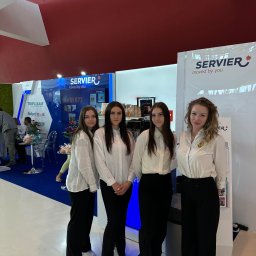 Eventship Weronika Pietruszka - Cztery kobiety w białych koszulach i czarnych spodniach stoją na tle stoiska targowego z logo firmy Servier, widoczny ekspres do kawy i inne elementy stoiska.