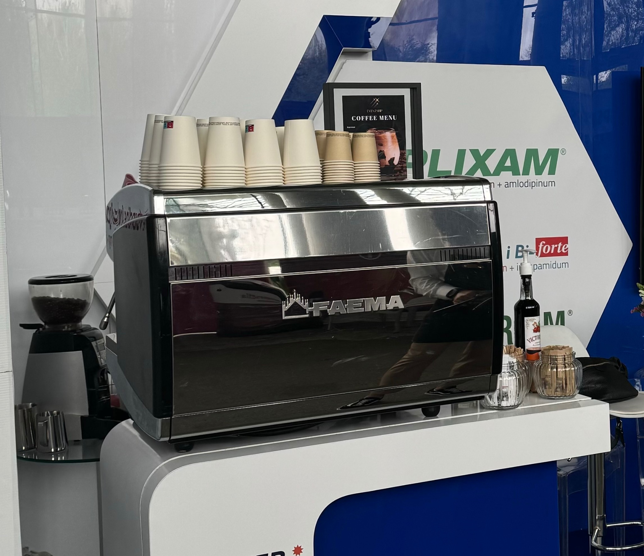 Profesjonalny ekspres do kawy marki Faema z zapasem papierowych kubków, młynek do kawy i dodatki na blacie w otoczeniu materiałów reklamowych.