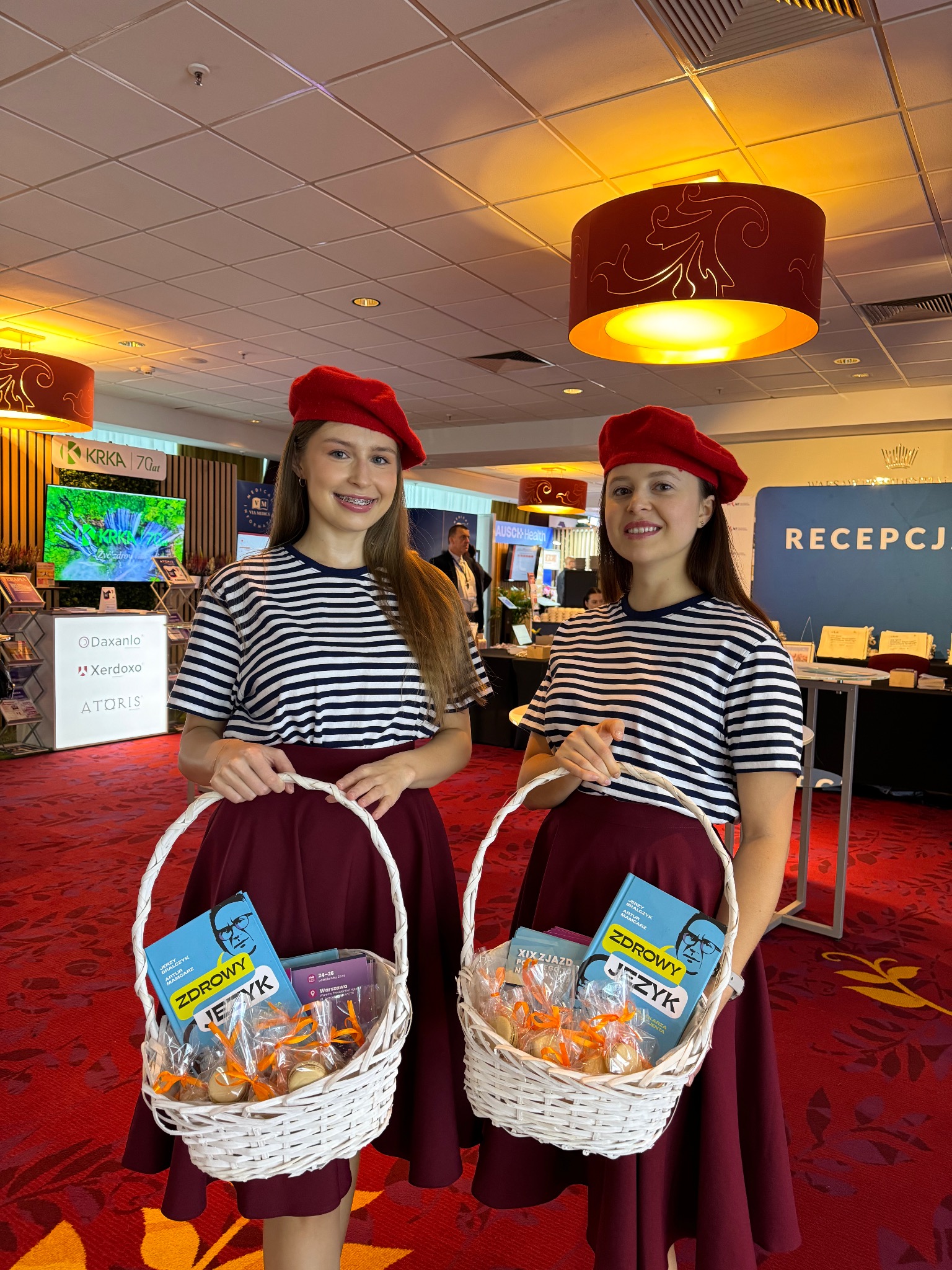 Dwie uśmiechnięte kobiety w pasiaste koszulki i czerwonych beretach prezentują koszyki z książkami i słodyczami na tle stoisk targowych z logo firm i napisem 'Recepcja'.