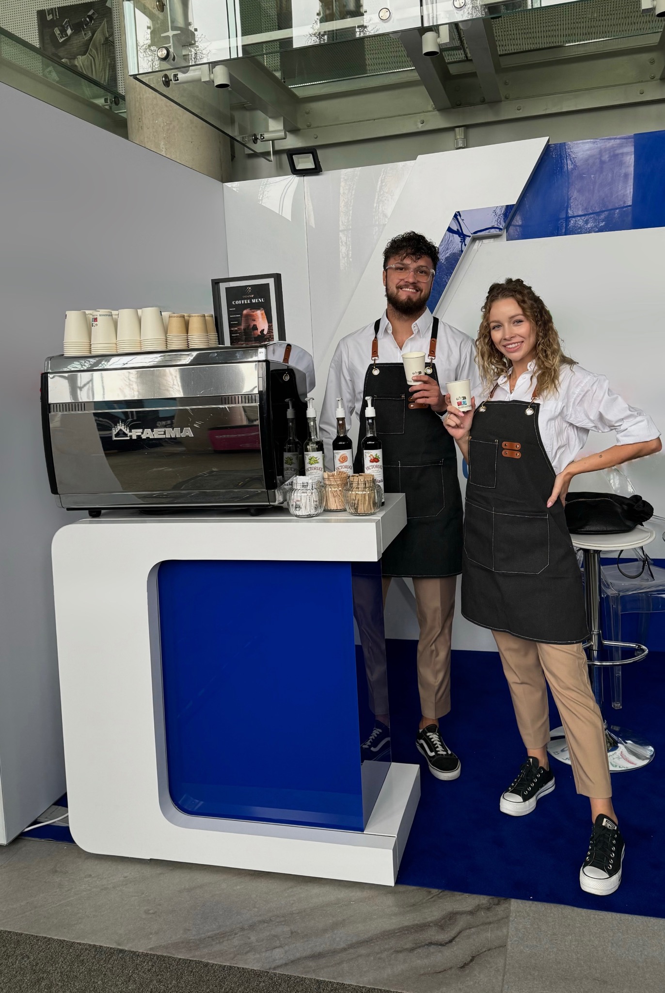 Uśmiechnięci barista i baristka w czarnych fartuchach, trzymający kubki z kawą obok profesjonalnego ekspresu Faema i menu kawowe w tle, na stoisku o nowoczesnym designie z niebieskimi akcentami.