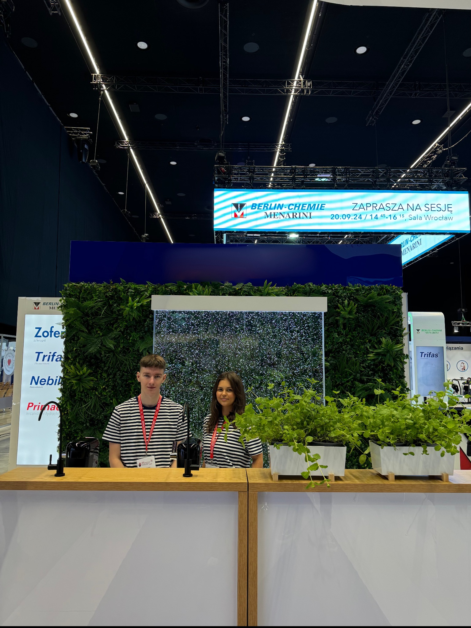 Stoisko targowe firmy farmaceutycznej Berlin-Chemie Menarini z dwoma pracownikami w pasiastej odzieży, dekoracją z zielonej ściany i fontanną z bąbelkami, oświetlone banerem LED zapraszającym...