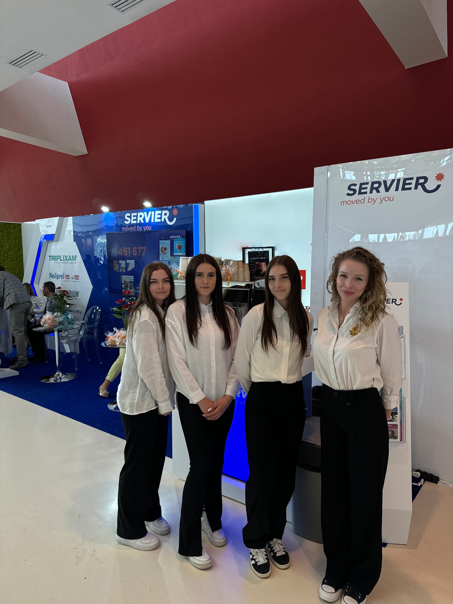 Cztery kobiety w białych koszulach i czarnych spodniach stoją na tle stoiska targowego z logo firmy Servier, widoczny ekspres do kawy i inne elementy stoiska.