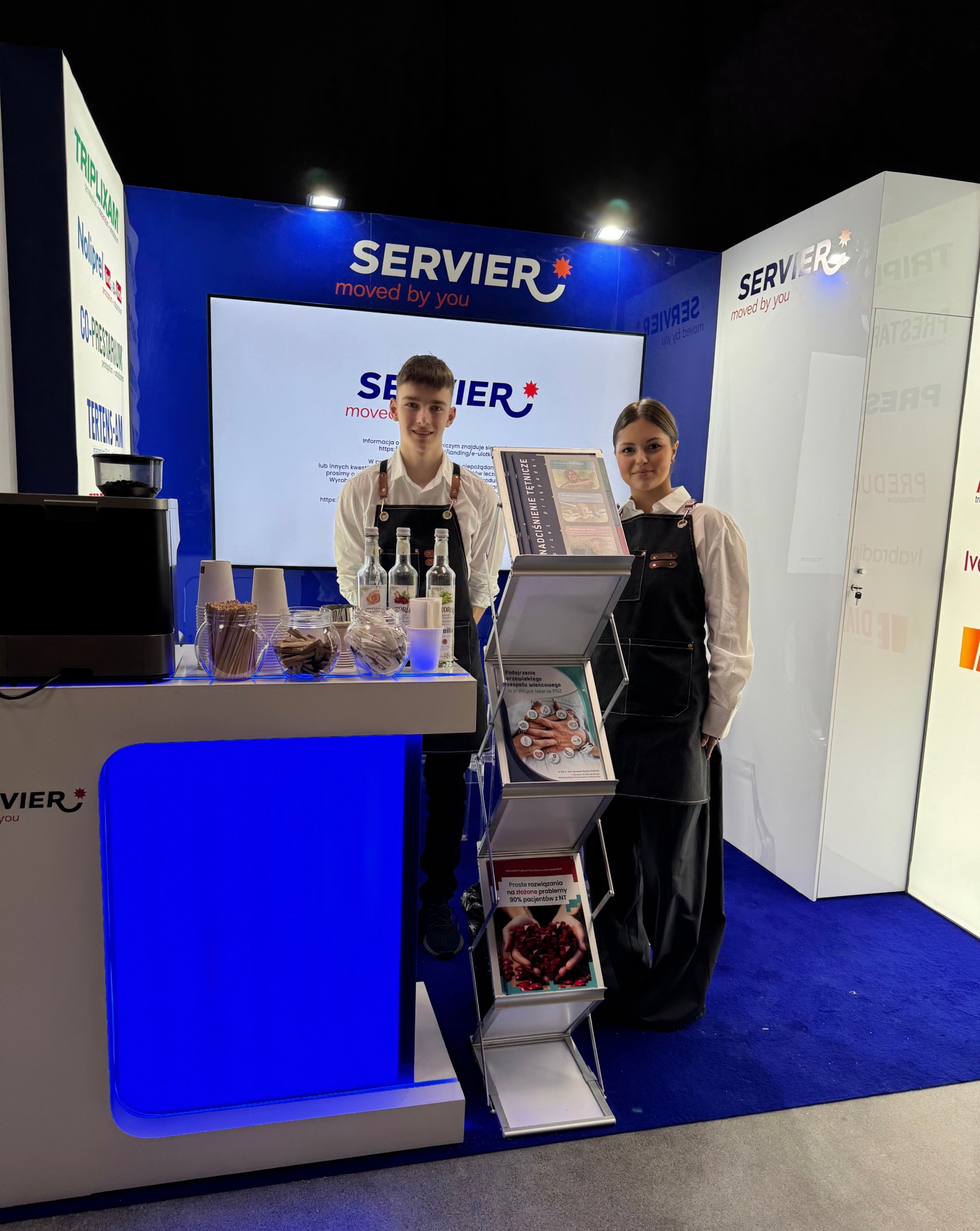 Stoisko firmowe na targach z dwójką osób w fartuchach, ekspres do kawy, materiały informacyjne i logo firmy w tle.