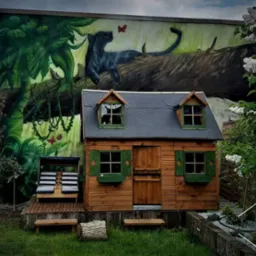Ściana budynku z muralem przedstawiającym czarną panterę w dżungli, obok drewniany domek dla dzieci z zielonymi okiennicami.