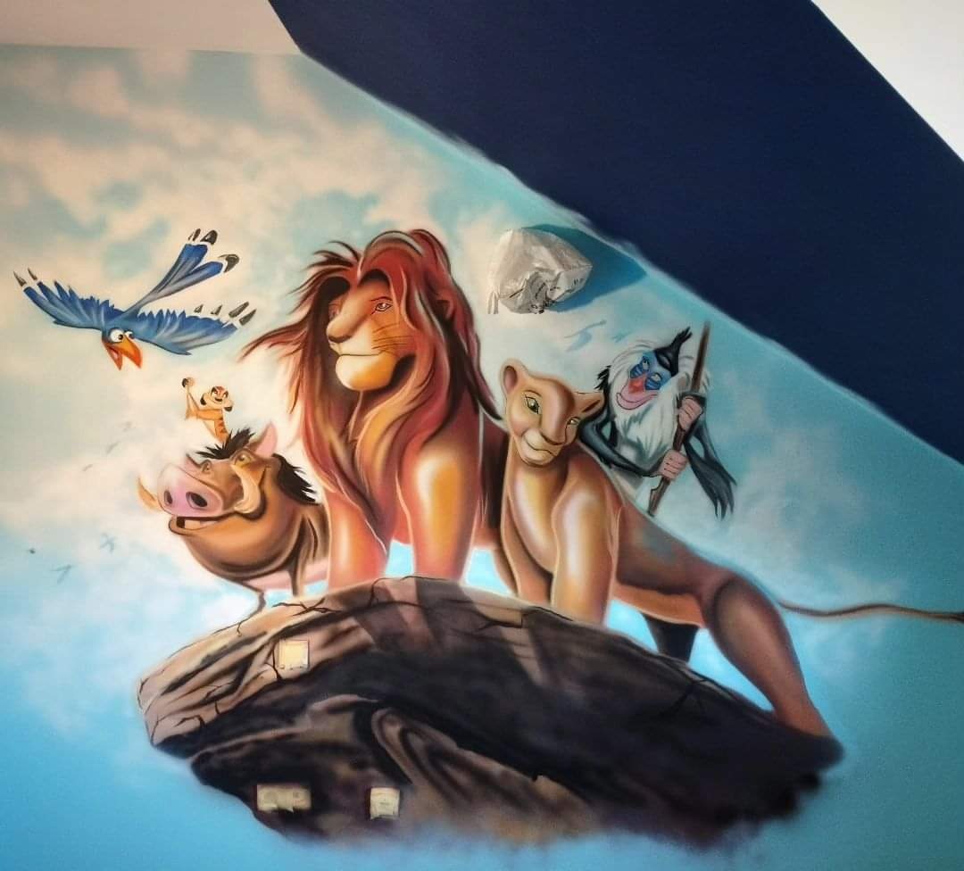 Mural na ścianie pokoju dziecięcego przedstawiający postacie z Króla Lwa: Simbę, Nalę, Rafikiego, Pumbę, Timona i Zazu na tle błękitnego nieba z chmurami, fragmenty taśmy malarskiej widoczne na górze