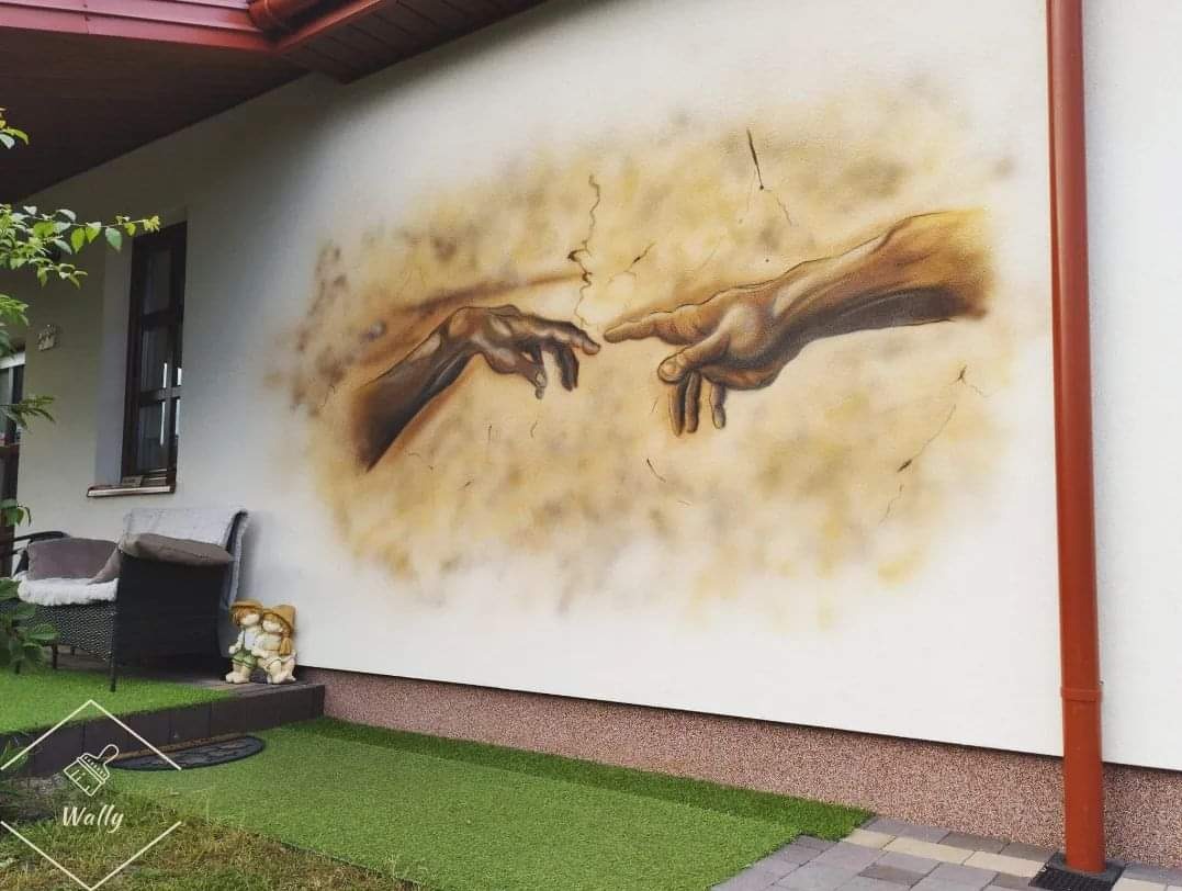 Zewnętrzny mural na ścianie domu, przedstawiający zbliżające się dłonie, nawiązujący do 'Stworzenia Adama' Michała Anioła, z widocznym fragmentem ogrodu i elementami architektury.