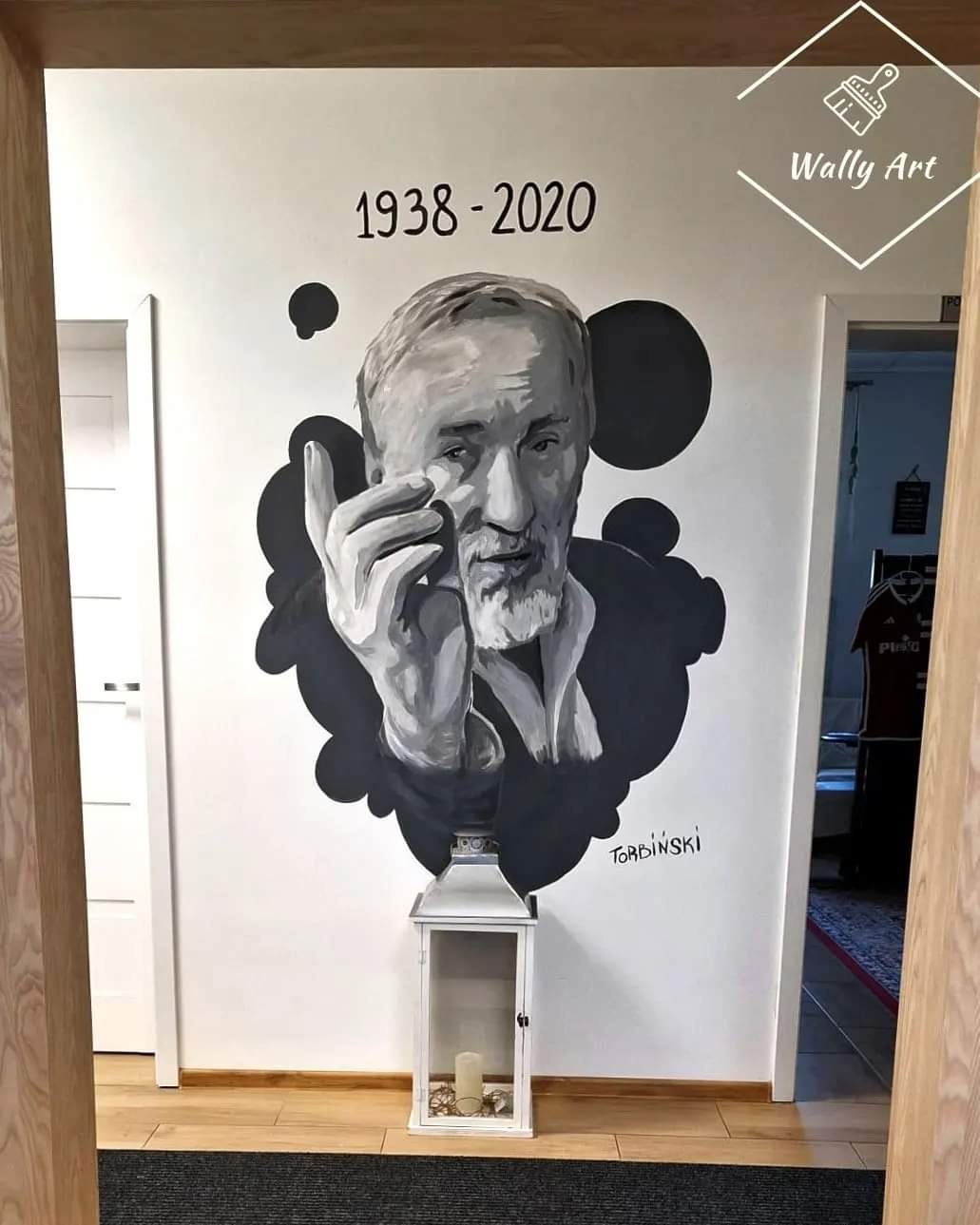 Czarno-biały portret malowany na ścianie przedstawiający mężczyznę z datami 1938-2020, poniżej napis 'Torbinski', na podłodze biały lampion ze świecą, widok z korytarza z dwiema parami drzwi.