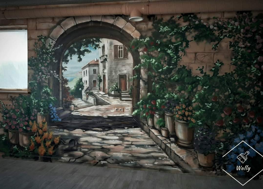Iluzjonistyczne malowidło ścienne przedstawiające toskańską uliczkę z kamienną bramą, kwiatami w donicach i budynkami w oddali, wykonane z efektem 3D na ścianie w pomieszczeniu.