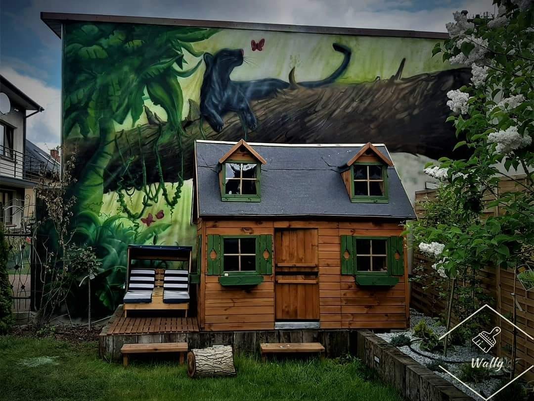 Ściana budynku z muralem przedstawiającym czarną panterę w dżungli, obok drewniany domek dla dzieci z zielonymi okiennicami.