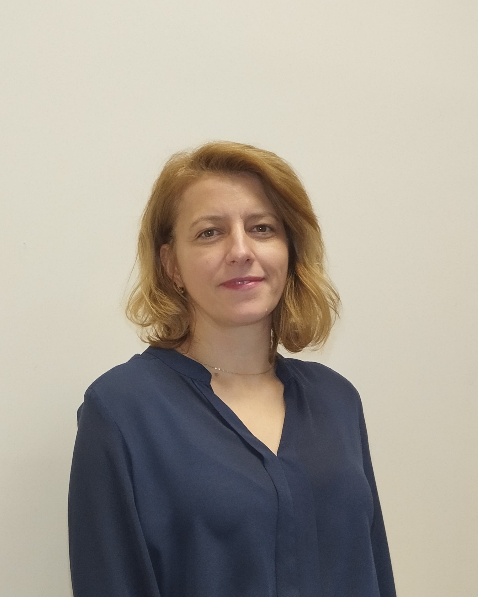 Portret kobiety z krótkimi blond włosami, ubranej w granatową bluzkę na tle jasnej ściany.