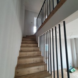 Industrial Story - Drewniane schody z czarną metalową balustradą prowadzące na antresolę w surowym wnętrzu, widoczne ślady po pracach wykończeniowych na ścianach.