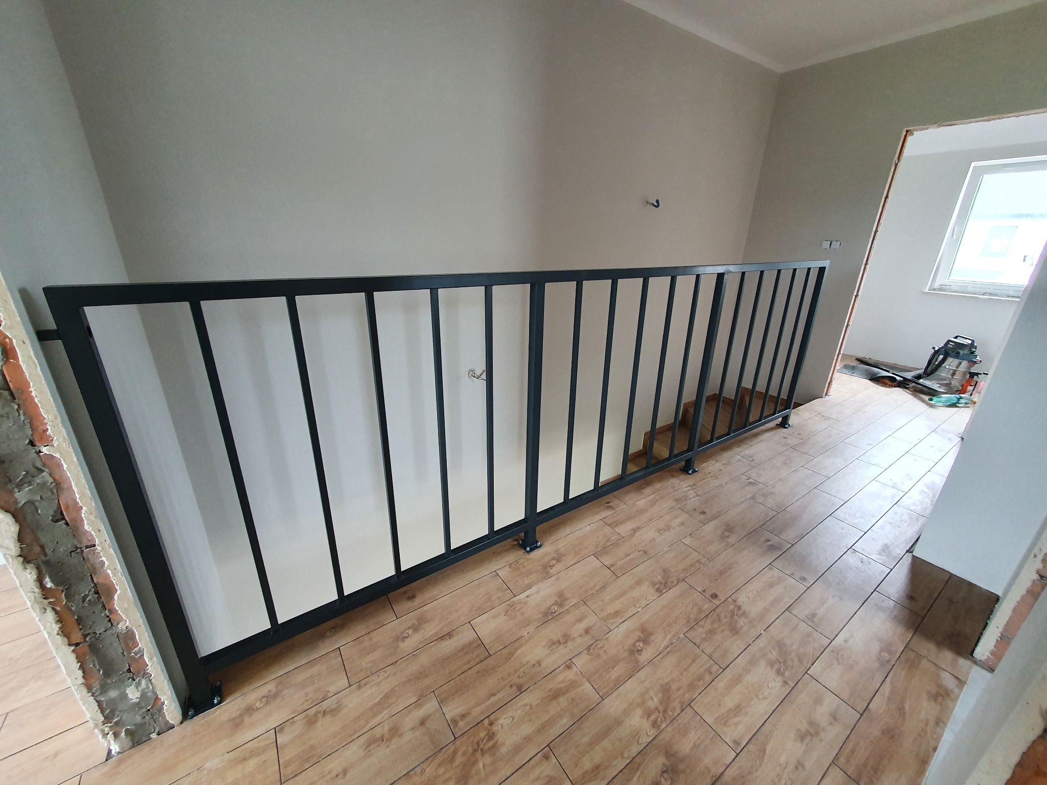 Nowoczesna, czarna balustrada metalowa z pionowymi szczeblami zamontowana wewnątrz budynku, widoczne surowe wykończenie ścian i drewnopodobne płytki na podłodze.