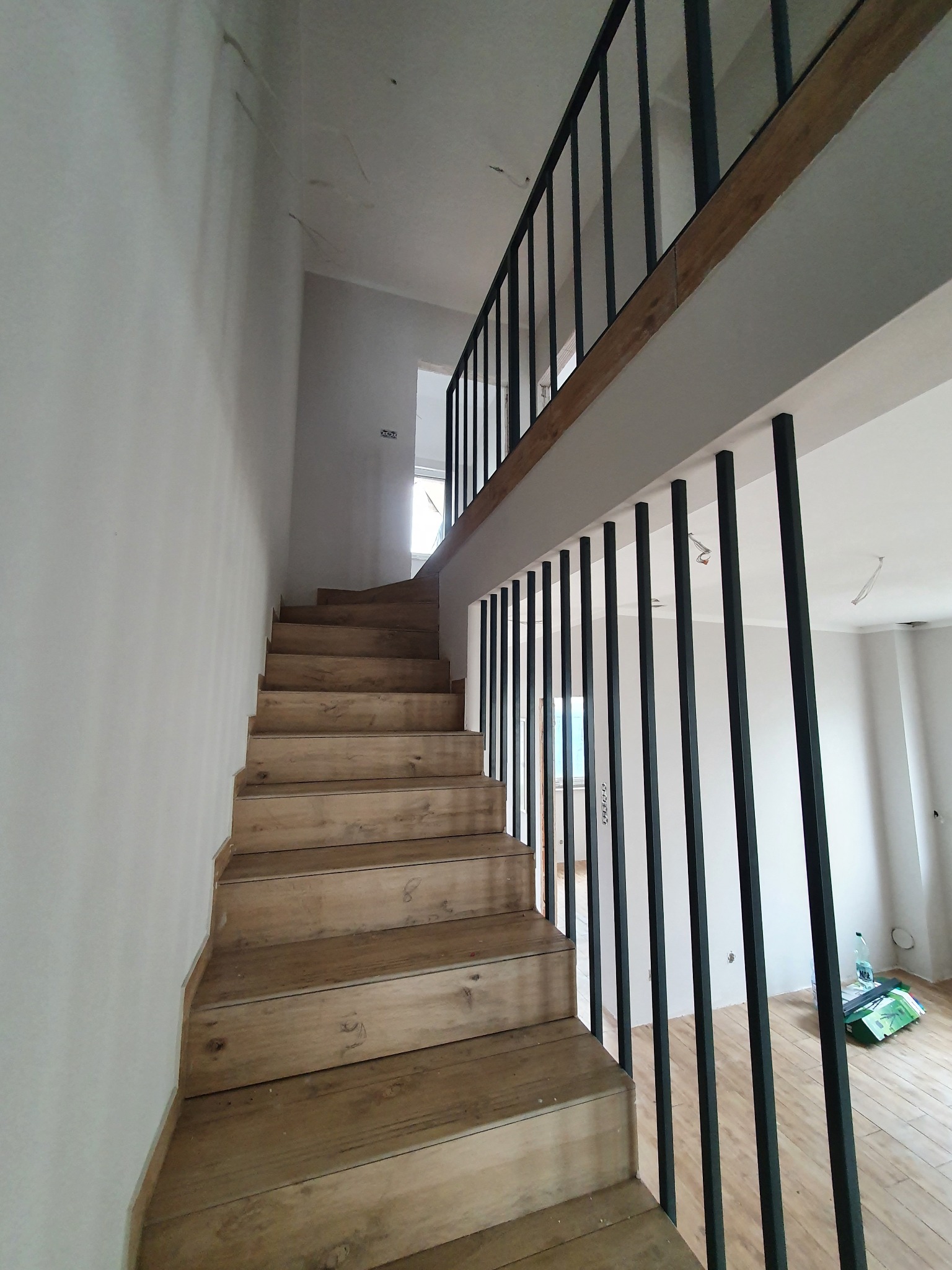 Drewniane schody z czarną metalową balustradą prowadzące na antresolę w surowym wnętrzu, widoczne ślady po pracach wykończeniowych na ścianach.