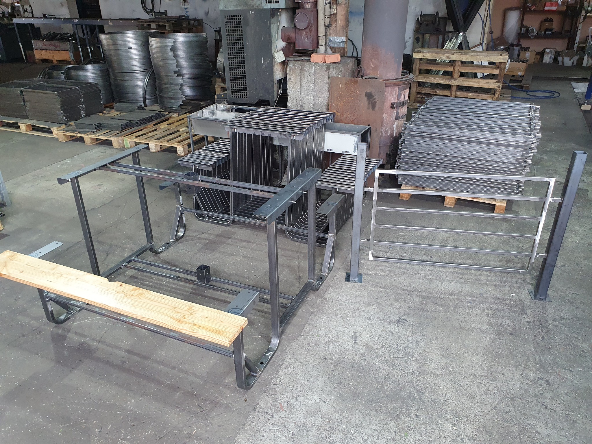 Warsztatowe ujęcie metalowych konstrukcji: rama ławki z drewnianym siedziskiem, kratownica oraz elementy ogrodzenia w surowym stanie, ułożone na betonowej posadzce, w tle stosy metalowych...