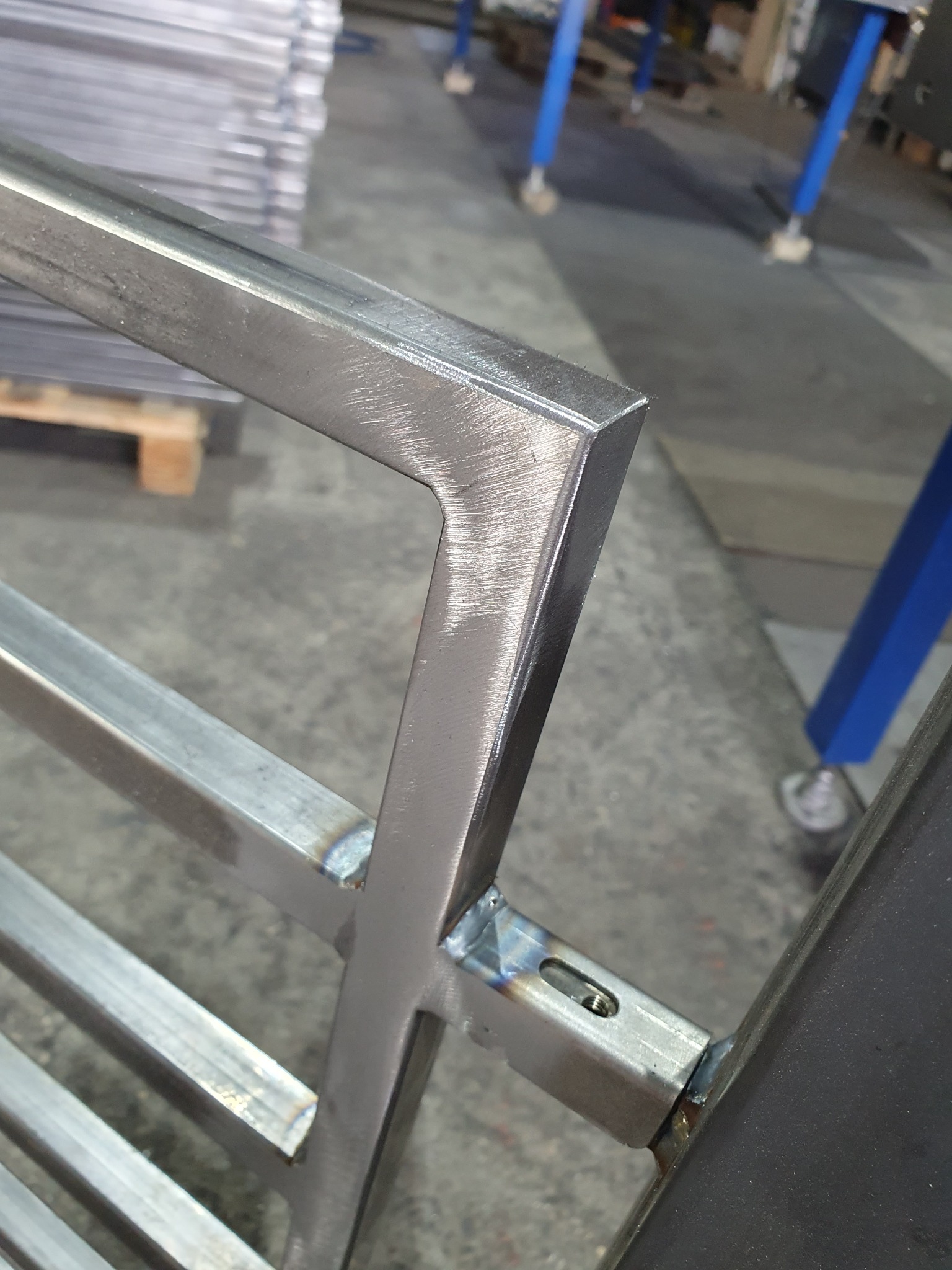 Fragment konstrukcji metalowej z profili zamkniętych, świeżo po spawaniu, widoczne spawy i ślady obróbki szlifierskiej, w tle elementy warsztatu.