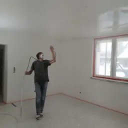 Mężczyzna w masce maluje sufit w pustym pokoju za pomocą pistoletu natryskowego; folia ochronna na podłodze i oknach.