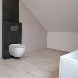 Nowoczesna toaleta podwieszana z szarą obudową i chromowanym przyciskiem spłukiwania na tle beżowych płytek podłogowych i ściany w skosie, widoczny fragment białej wanny.