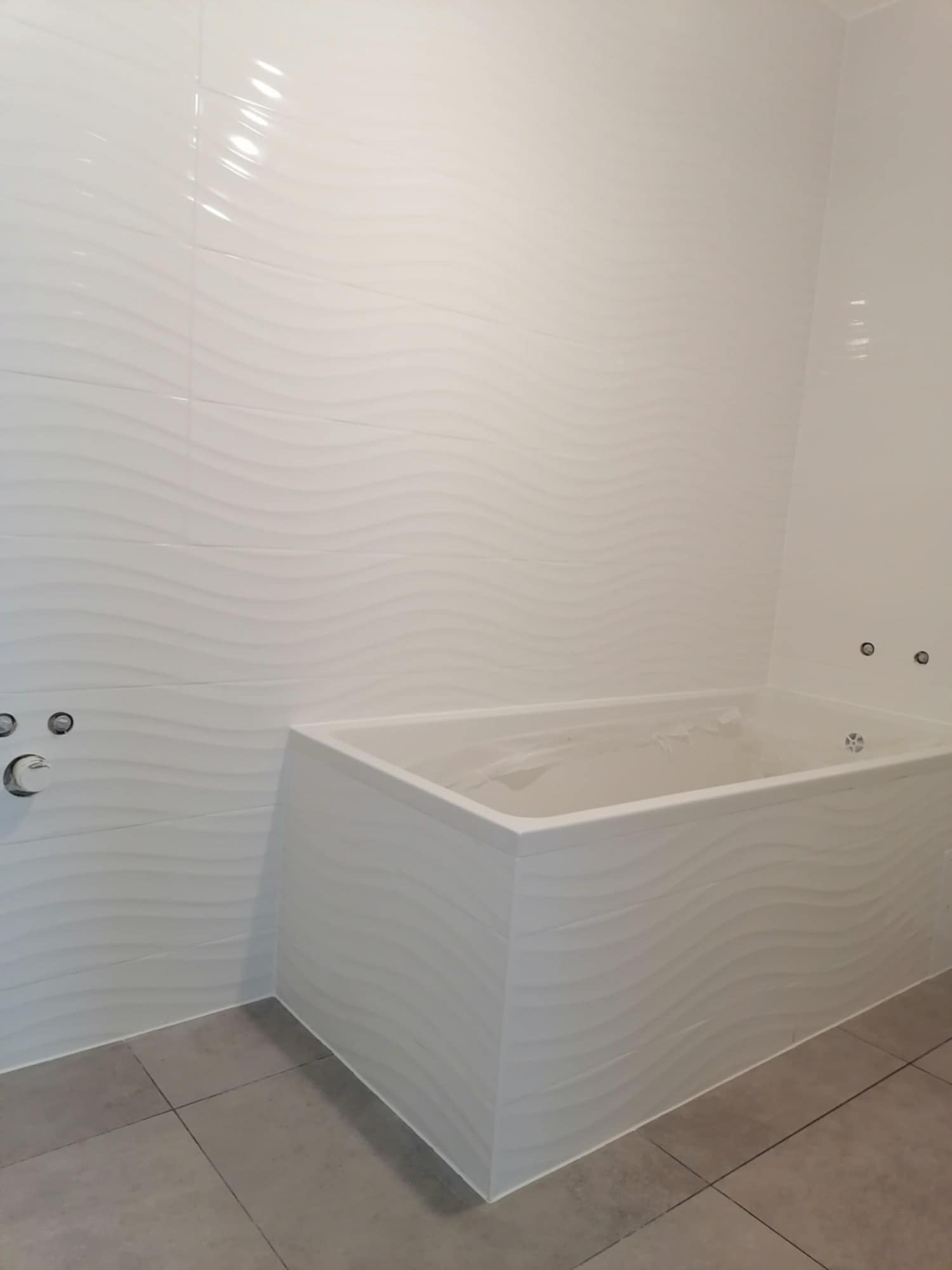 Nowoczesna łazienka z białą wanną z hydromasażem, wykończona płytkami ściennymi w falisty wzór i szarą podłogą z płytek.