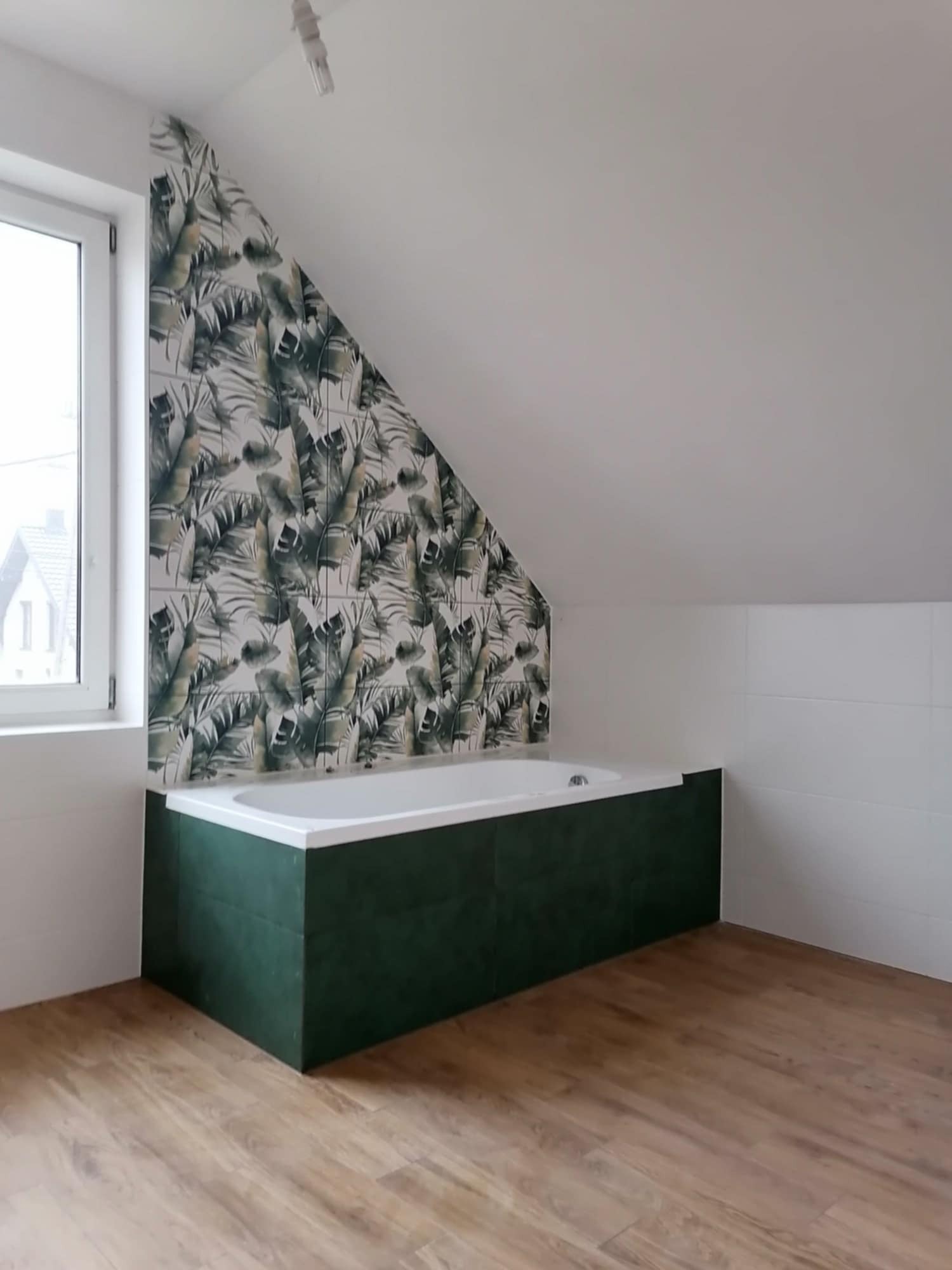 Biała wanna obudowana ciemnozielonymi płytkami w łazience na poddaszu, ściana pokryta płytkami z motywem liści, drewnopodobna podłoga, białe płytki na ścianie.