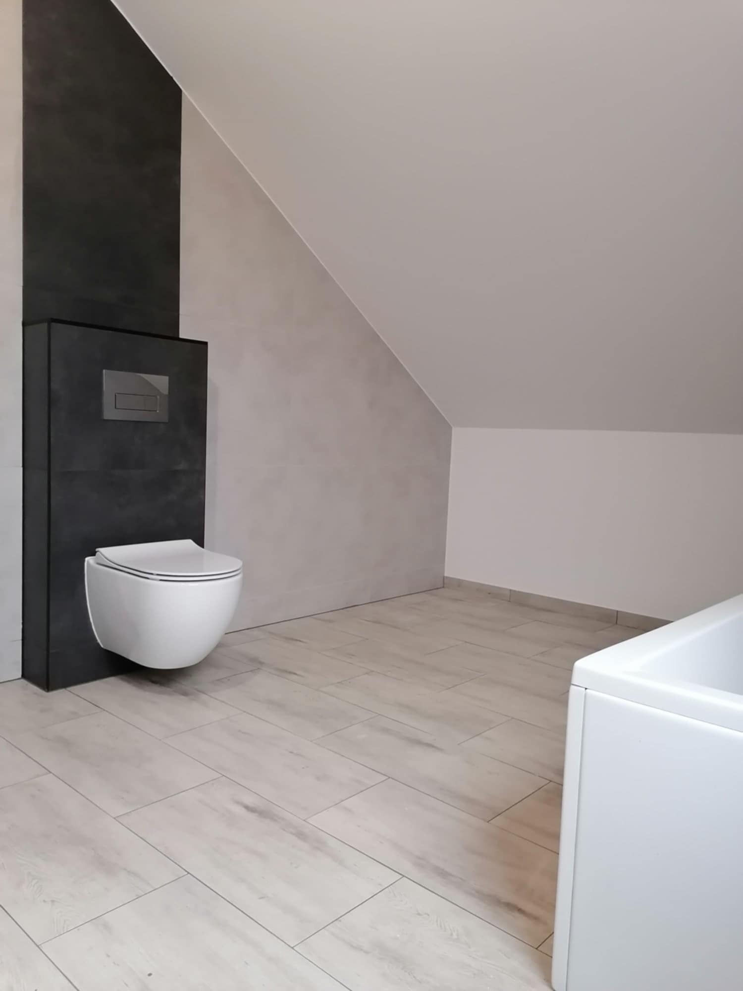 Nowoczesna toaleta podwieszana z szarą obudową i chromowanym przyciskiem spłukiwania na tle beżowych płytek podłogowych i ściany w skosie, widoczny fragment białej wanny.