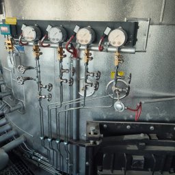 NORSU TECHNOLOGYS SPÓŁKA KOMANDYTOWA - Metalowa instalacja przemysłowa z manometrami, zaworami i rurami, zamontowana na teksturowanej, metalowej ścianie w hali.