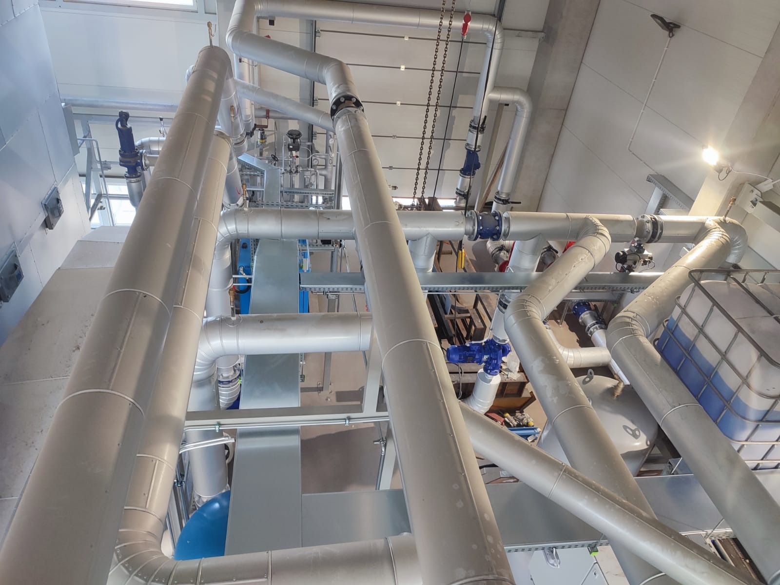 Rozbudowany system rur przemysłowych z zaworami i zbiornikami, widok z dołu, oświetlenie górne.