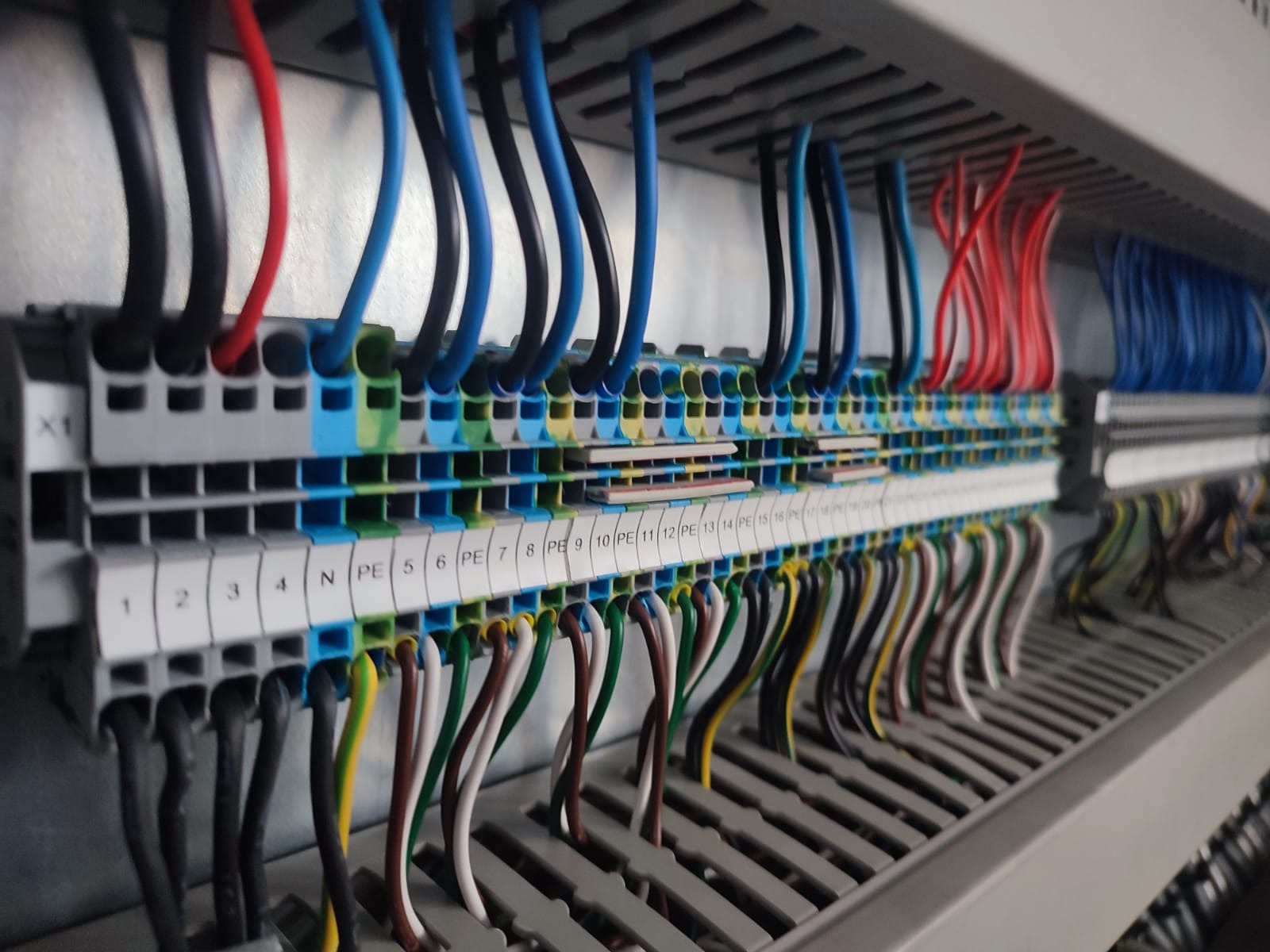 Rozdzielnica elektryczna z widocznymi przewodami o różnych kolorach (niebieski, czerwony, czarny, zielono-żółty) podłączonymi do listew zaciskowych z numeracją.