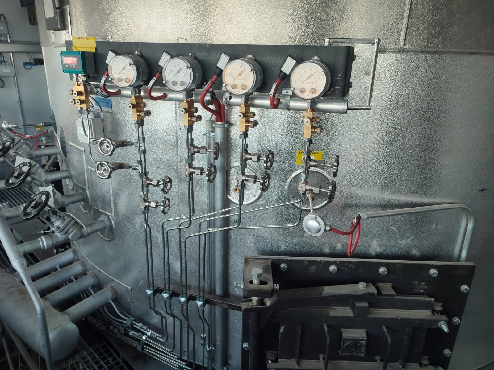 Metalowa instalacja przemysłowa z manometrami, zaworami i rurami, zamontowana na teksturowanej, metalowej ścianie w hali.