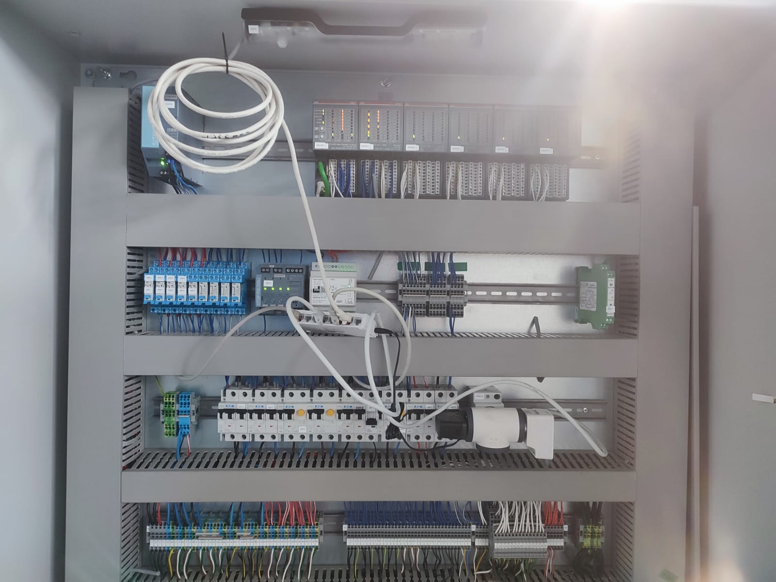 Wnętrze szafy sterowniczej z modułami PLC, przekaźnikami, zasilaczami i okablowaniem, widok z góry, oświetlenie sufitowe.