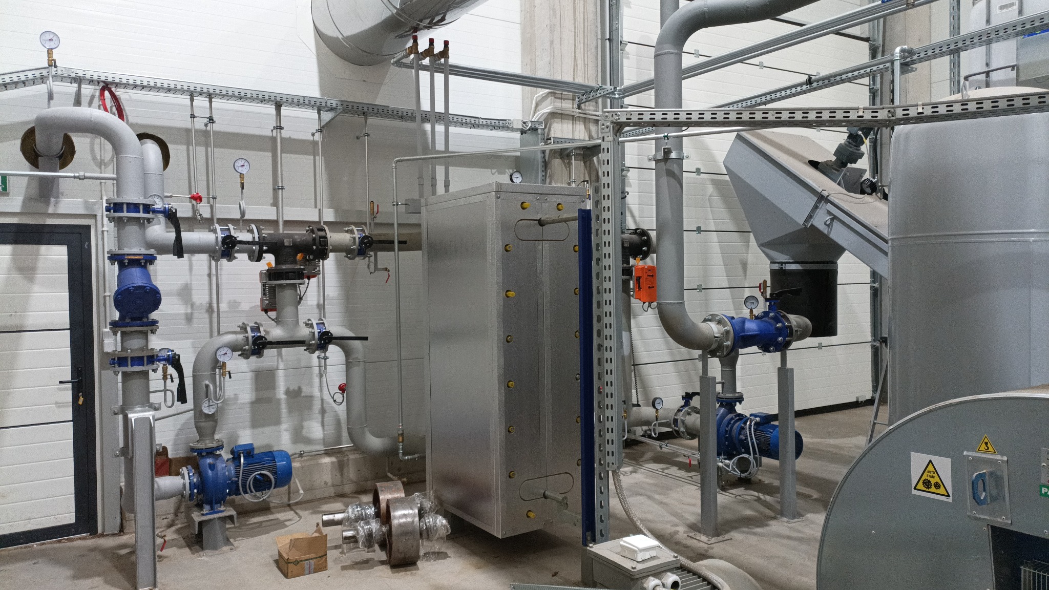 Wnętrze hali z zainstalowaną, skomplikowaną instalacją przemysłową z rur, zaworów, pomp w kolorze niebieskim i srebrnym, oraz zbiornika. Widoczne manometry i elementy mocujące.