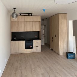 PAVLO YAKOVENKO IN-ARK - Jasna, drewnopodobna zabudowa kuchenna z czarnym blatem i sprzętami AGD. Na podłodze panele, w tle szafa i narzędzia. Lampa wisząca w folii ochronnej.