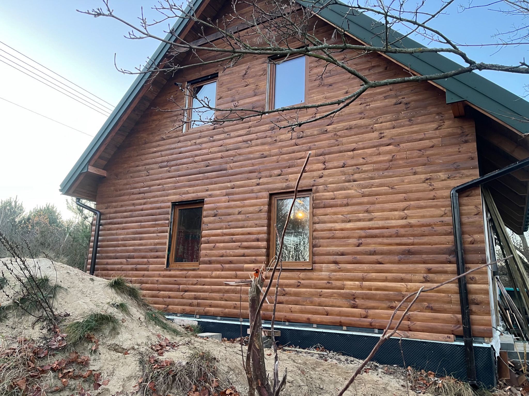 Drewniany dom z bali z zielonym dachem i widocznymi oknami. Świeżo wybudowany, z piaskiem i resztkami budowy wokół. Widok z dołu, podkreślający strukturę drewna.