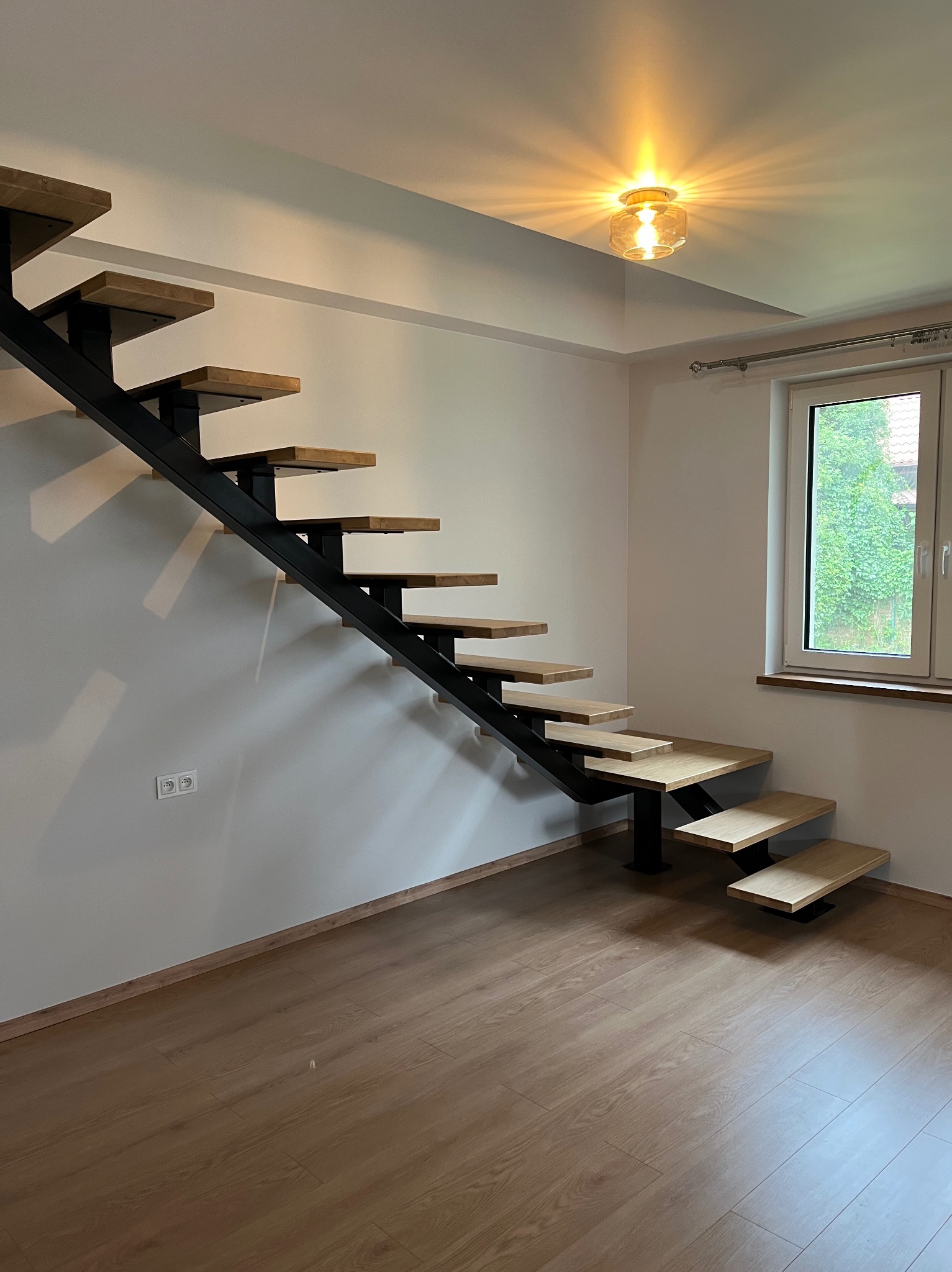 Nowoczesne schody wiszące z drewnianymi stopniami i czarną metalową konstrukcją, oświetlone ciepłym światłem lampy sufitowej, na tle białej ściany i drewnianej podłogi.