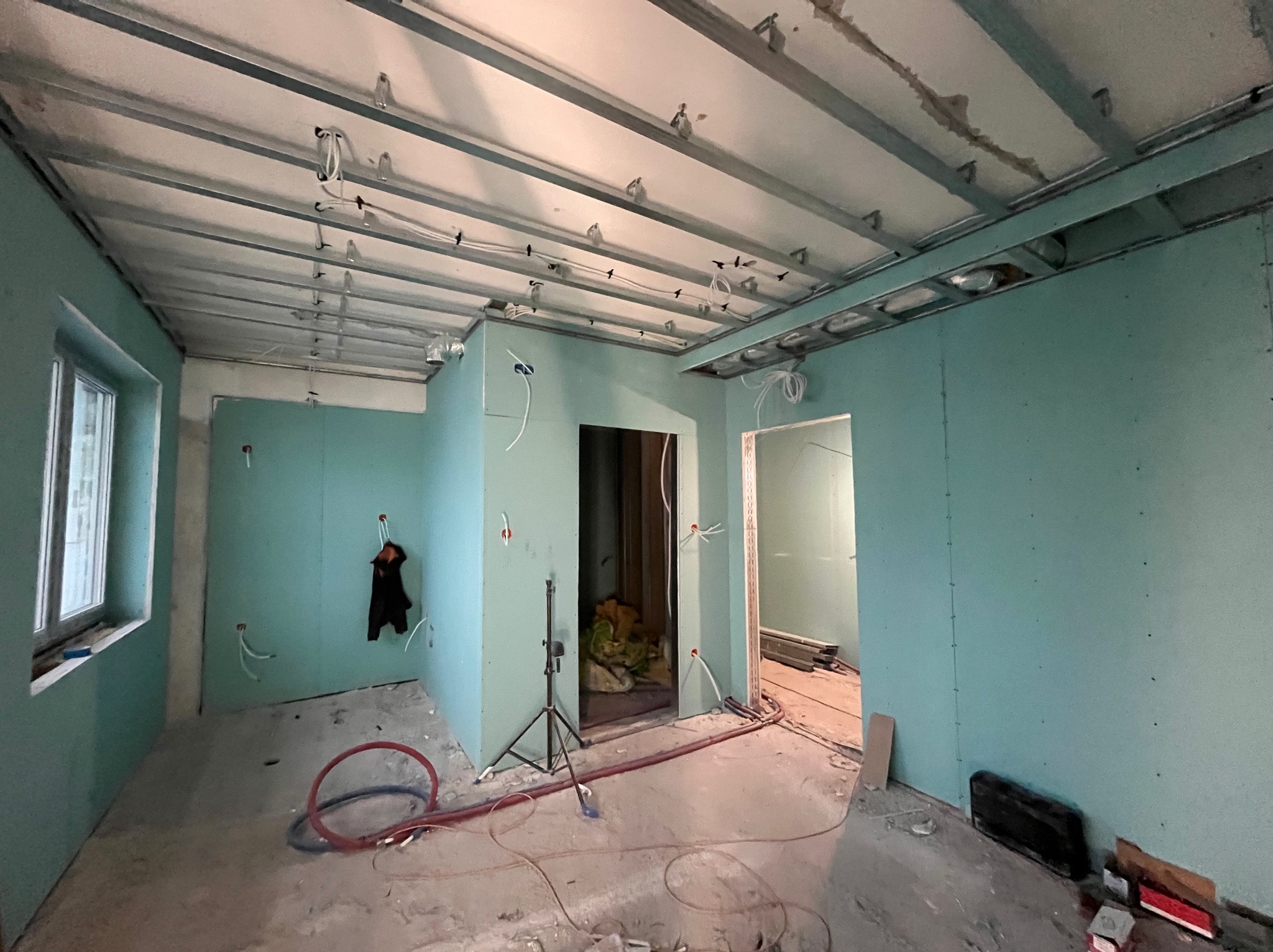 Wnętrze w trakcie remontu: ściany z płyt karton-gips w odcieniach turkusu, metalowa konstrukcja sufitu podwieszanego z instalacją elektryczną, widoczne kable i narzędzia.