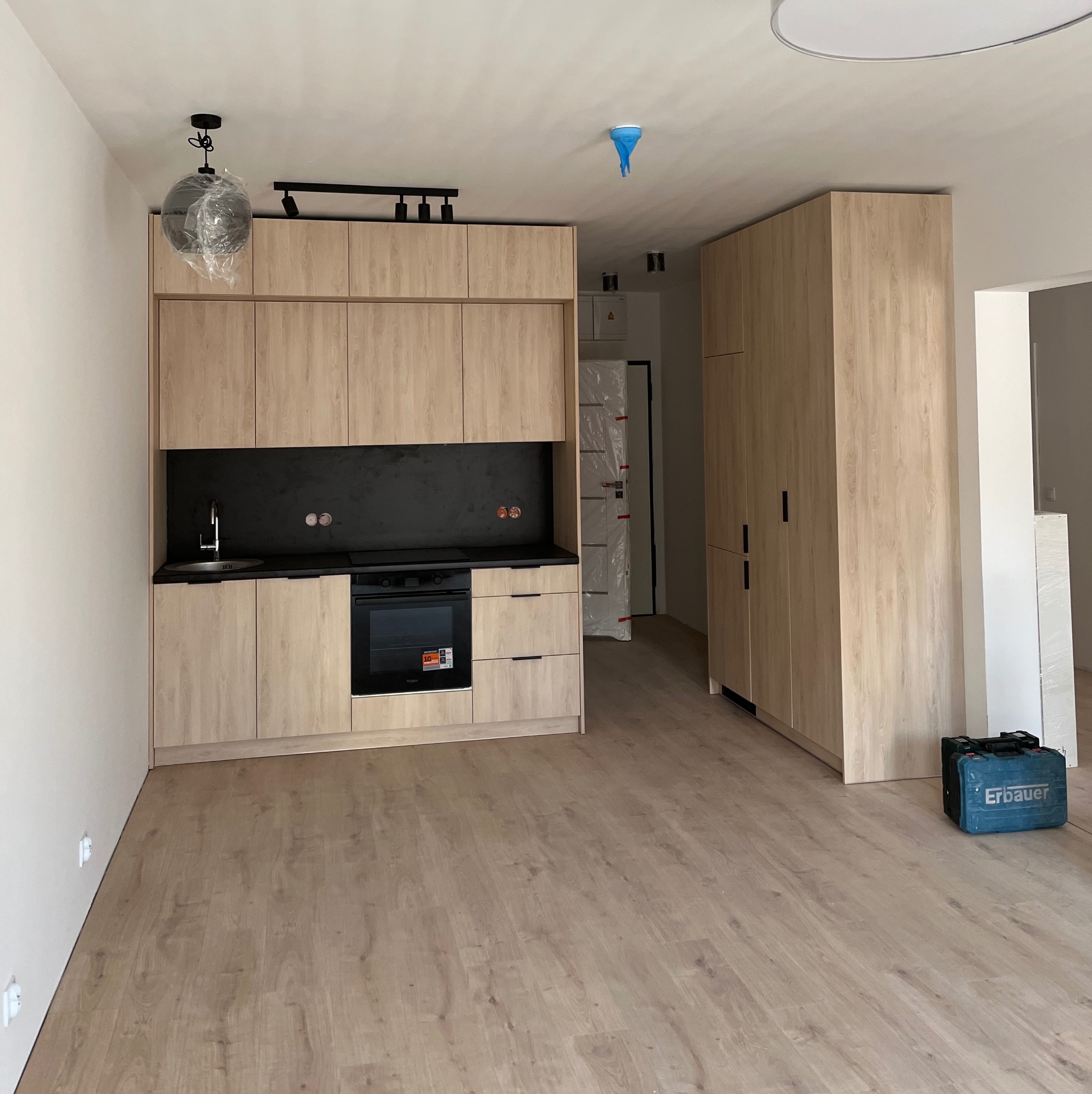 Jasna, drewnopodobna zabudowa kuchenna z czarnym blatem i sprzętami AGD. Na podłodze panele, w tle szafa i narzędzia. Lampa wisząca w folii ochronnej.
